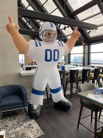 Inflatable giant football player mascot in white-and-blue uniform (00) inside a rooftop lounge with panoramic city skyline views