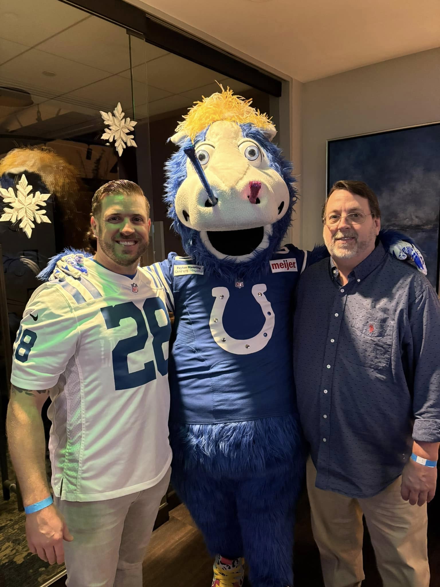 Two men posing with a blue horse football mascot in a sports lounge — one man wearing a white #28 jersey, festive snowflake window decals in background.