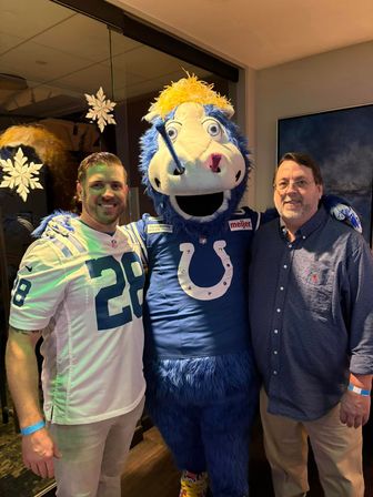 Two men posing with a blue horse football mascot in a sports lounge — one man wearing a white #28 jersey, festive snowflake window decals in background.