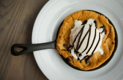 Warm chocolate chip skillet cookie a la mode in a small cast-iron pan, topped with a scoop of vanilla ice cream and chocolate drizzle, served on a white plate over a wooden table.