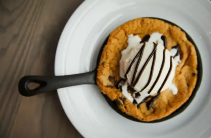 Warm chocolate chip skillet cookie a la mode in a small cast-iron pan, topped with a scoop of vanilla ice cream and chocolate drizzle, served on a white plate over a wooden table.