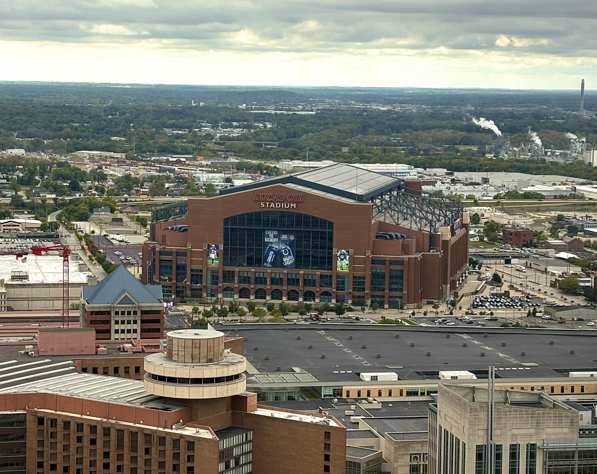 Aerial shot of a giant brick stadium with a glass façade and retractable roof rising above surrounding warehouses, parking lots and low-rise buildings under a moody cloudy sky.