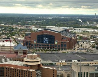 Aerial shot of a giant brick stadium with a glass façade and retractable roof rising above surrounding warehouses, parking lots and low-rise buildings under a moody cloudy sky.