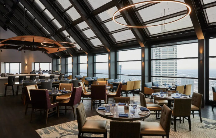 Stylish rooftop restaurant with glass atrium and floor-to-ceiling windows showcasing panoramic urban skyline views, round pendant lights, and neatly set dining tables.
