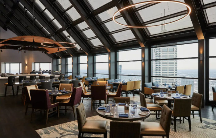 Stylish rooftop restaurant with glass atrium and floor-to-ceiling windows showcasing panoramic urban skyline views, round pendant lights, and neatly set dining tables.