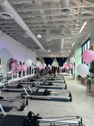 Light-filled Pilates reformer studio with rows of reformer machines, pink and white balloons along mirrors and windows, exposed white ductwork ceiling and a small group gathered near dark curtains.