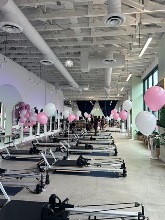 Light-filled Pilates reformer studio with rows of reformer machines, pink and white balloons along mirrors and windows, exposed white ductwork ceiling and a small group gathered near dark curtains.