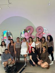 Smiling group in a bright modern pilates studio posing by a reformer with pink and white balloons and large pink '28' number balloons, arched mirrors reflecting a colorful mural.