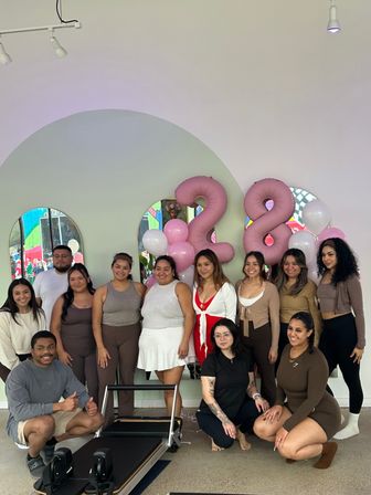 Smiling group in a bright modern pilates studio posing by a reformer with pink and white balloons and large pink '28' number balloons, arched mirrors reflecting a colorful mural.
