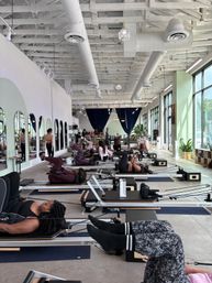 Bright modern Pilates studio with rows of reformer machines and a group reformer class; people in athletic wear exercising in a spacious, sunlit space with exposed white-beam ceiling, floor-to-ceiling windows and green-accented walls.