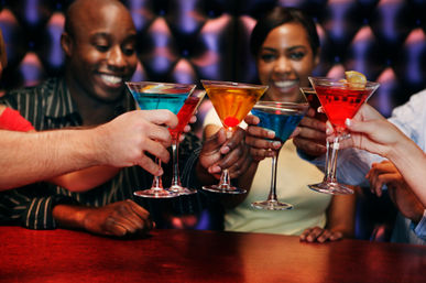 Smiling friends clinking colorful martini glasses at a lively bar — blue, red and amber cocktails in a celebratory toast, upbeat nightlife scene.