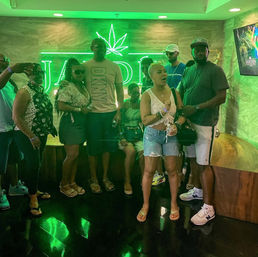 Group of friends posing in a neon green cannabis lounge under a glowing leaf sign, wearing casual summer outfits with wooden seating and a reflective black floor.