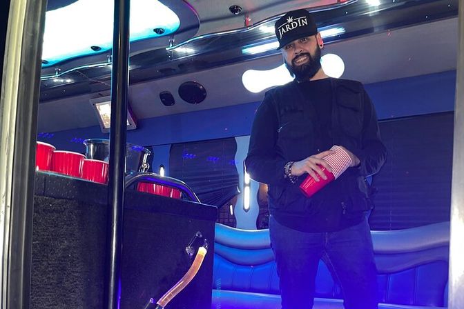 Man in black jacket and baseball cap holding stacked red plastic cups inside a blue-lit party bus with leather seating, a metal pole, and neon ceiling lights.