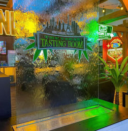Cascading water wall inside a Las Vegas tasting room, featuring a green cannabis-leaf logo, colorful neon signs and reflections with a potted plant nearby.