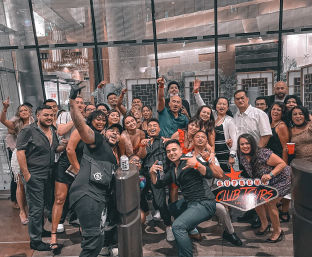 Energetic group of friends posing in front of glass-front venue in downtown Las Vegas nightlife, smiling, holding drinks and making celebratory gestures.