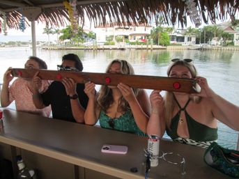 Four people taking synchronized shots from a long wooden communal shot board under a thatched tiki roof at a waterfront bar overlooking a canal with palm trees and waterfront homes.