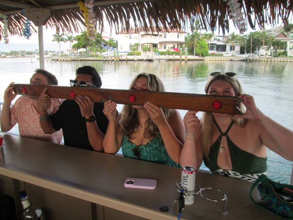 Four people taking synchronized shots from a long wooden communal shot board under a thatched tiki roof at a waterfront bar overlooking a canal with palm trees and waterfront homes.