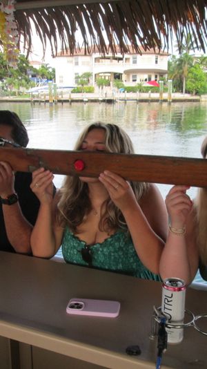 Four people taking synchronized shots from a long wooden communal shot board under a thatched tiki roof at a waterfront bar overlooking a canal with palm trees and waterfront homes.