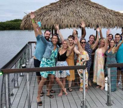 Happy group of adults waving under a thatched tiki hut on a wooden waterfront dock with calm water and mangrove shoreline — casual tropical vacation gathering