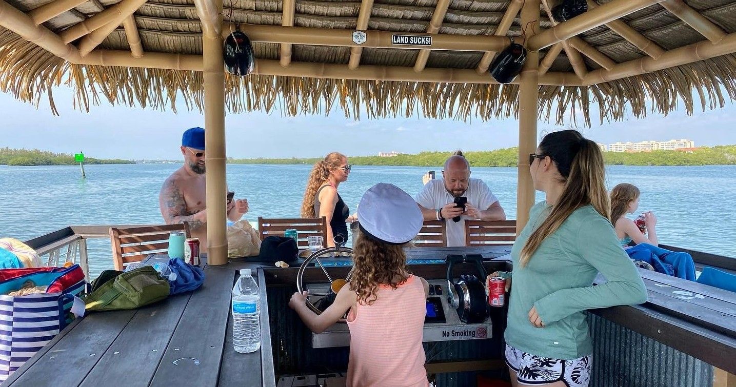 Child wearing a captain's hat steers a tiki-roofed boat while family and friends relax under a thatched hut on a sunny coastal inlet with calm blue water and mangroves.
