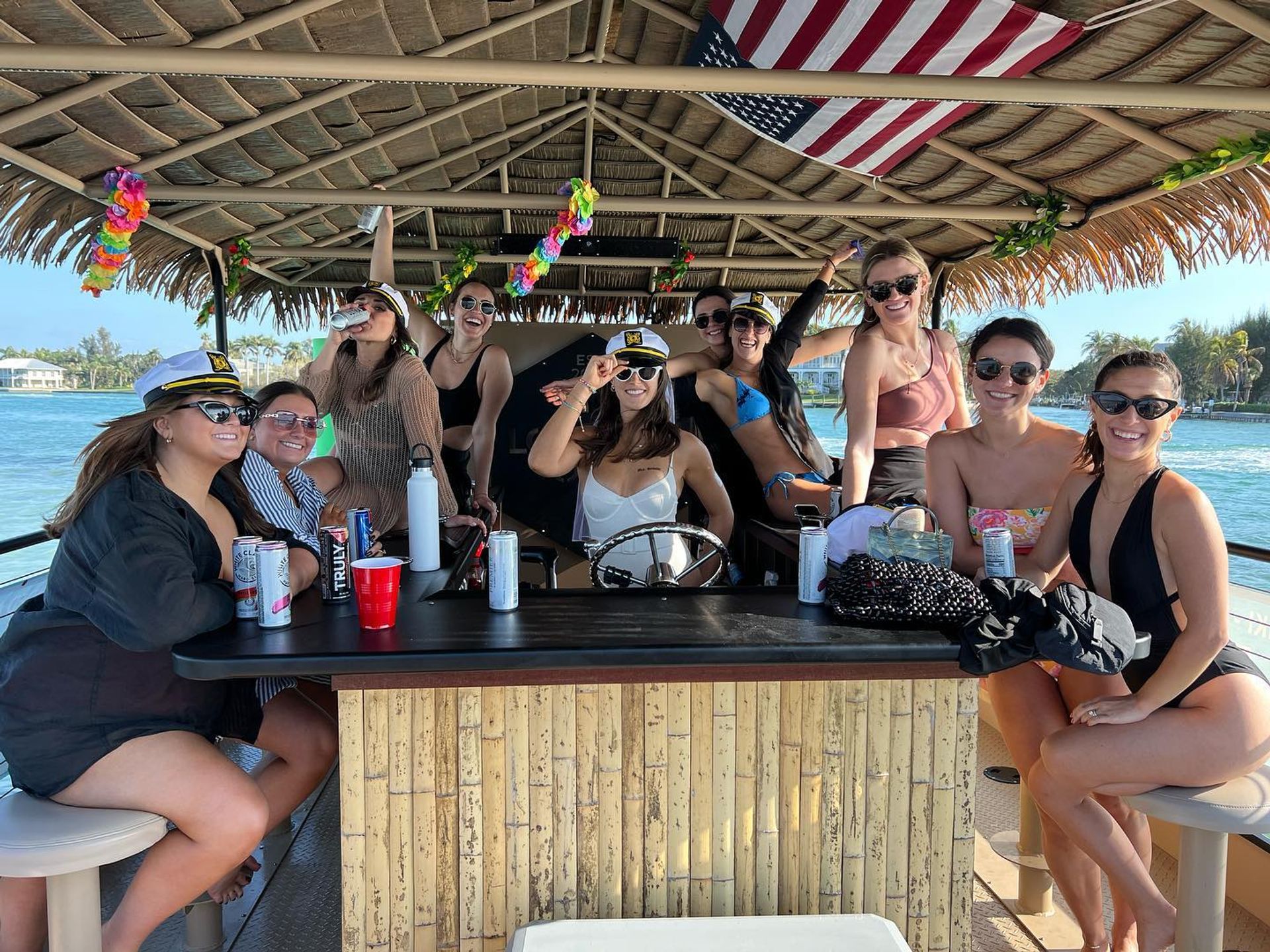 Group of smiling women in swimsuits and captain hats enjoying a sunny tiki-style pontoon boat party on turquoise coastal waters with palm trees and an American flag overhead