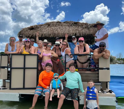 Smiling multigenerational group in swimwear on a tiki-roof pontoon boat in a sunny coastal bay, with kids in bright life jackets on the swim platform and waterfront homes and palm trees in the background.
