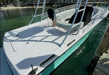Sunlit center-console boat with white deck and seafoam-green hull, stainless T-top frame and cushioned bench, moored at a wooden marina dock over clear green water.