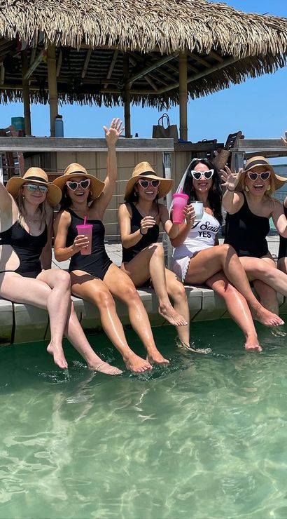 Friends in matching black swimsuits and straw hats sitting on a thatched-tiki dock over clear turquoise water, feet in the sea and holding bright pink cups on a sunny beach getaway.