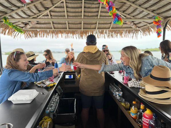 Friends sharing drinks on a tiki-thatched party boat bar, reaching for shot cups across the counter with water and shoreline visible in the background.