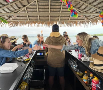 Friends sharing drinks on a tiki-thatched party boat bar, reaching for shot cups across the counter with water and shoreline visible in the background.