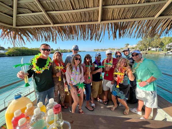 Cheerful group wearing colorful leis and sunglasses, holding palm-shaped tropical drinks under a thatched tiki roof on a sunny waterfront boat cruise near a marina.