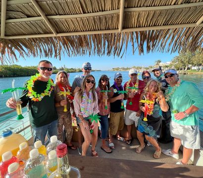 Cheerful group wearing colorful leis and sunglasses, holding palm-shaped tropical drinks under a thatched tiki roof on a sunny waterfront boat cruise near a marina.