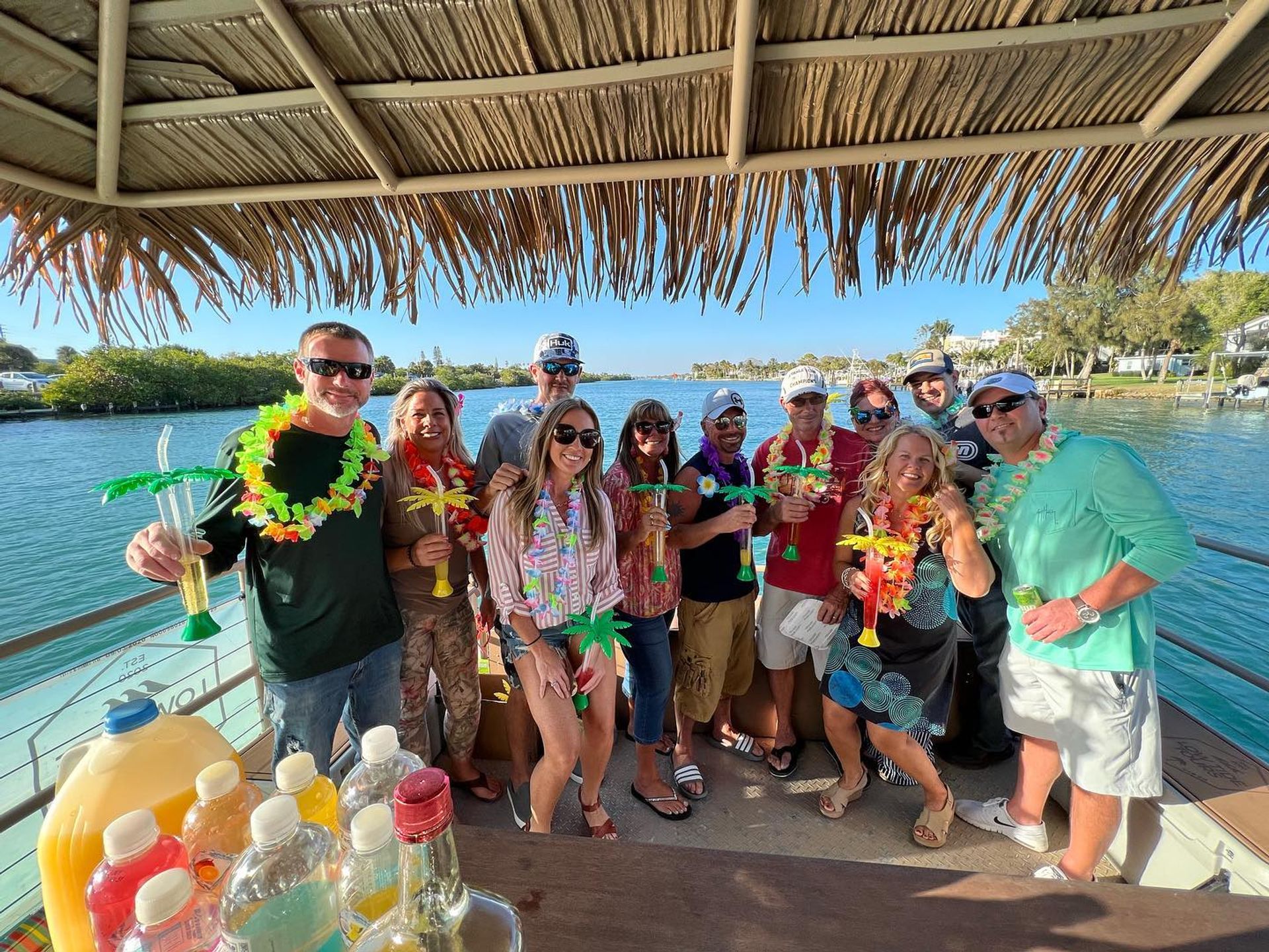 Cheerful group wearing colorful leis and sunglasses, holding palm-shaped tropical drinks under a thatched tiki roof on a sunny waterfront boat cruise near a marina.