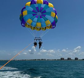 Two people parasailing over turquoise ocean beneath a colorful yellow, blue and purple parachute, towed by a boat with a coastal skyline and clear blue sky above.