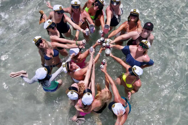 Aerial view of friends in swimsuits wearing captain hats standing in shallow beach water, forming a circle and raising cans for a sunny celebratory toast.