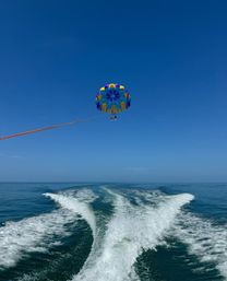 Colorful parasail carrying two riders above a calm blue ocean, trailing a boat's V-shaped wake under a clear sky.