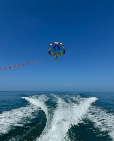 Colorful parasail carrying two riders above a calm blue ocean, trailing a boat's V-shaped wake under a clear sky.