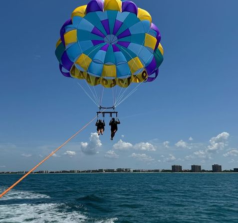 Two people parasailing beneath a bright blue, purple and yellow parachute over turquoise ocean with a distant coastal skyline and puffy clouds under a clear sunny sky
