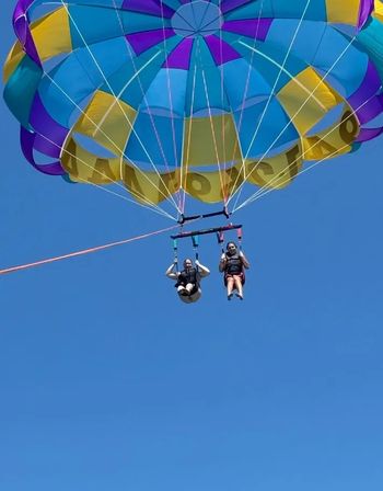 Tandem parasailing under a vibrant multicolored parachute (blue, yellow, purple) suspended by a towline against a clear blue sky