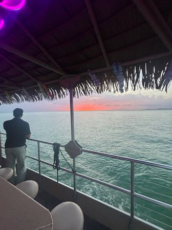 Silhouetted person leaning on the railing of a thatched-roof boat, watching a pink-orange tropical sunset over calm turquoise ocean
