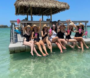 Seven friends in sun hats and heart-shaped sunglasses sitting on a tropical tiki dock, cheering with pink drink cups and dipping their feet in clear turquoise water under a bright blue sky.