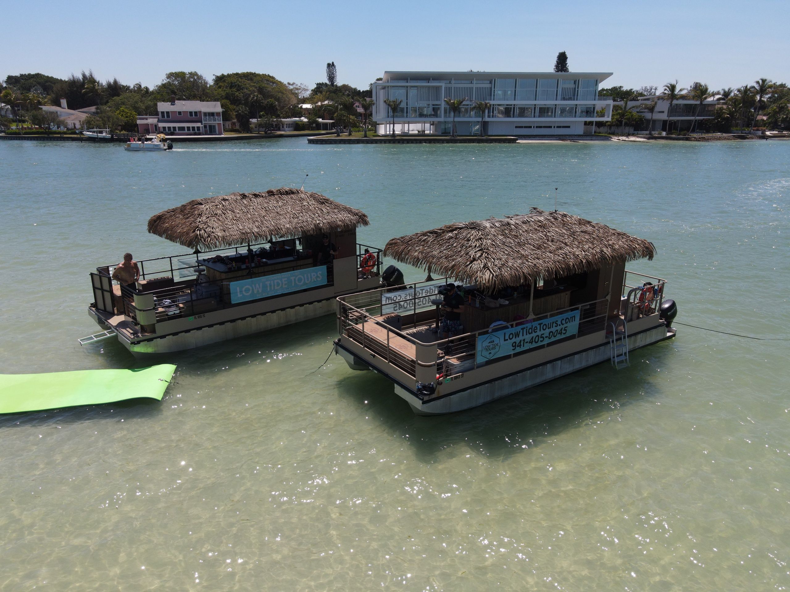 Two thatched-roof party pontoon boats anchored in clear shallow turquoise coastal bay, palm-lined waterfront homes in the background and a bright green floating mat nearby on a sunny day.