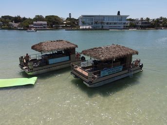 Two thatched-roof party pontoon boats anchored in clear shallow turquoise coastal bay, palm-lined waterfront homes in the background and a bright green floating mat nearby on a sunny day.