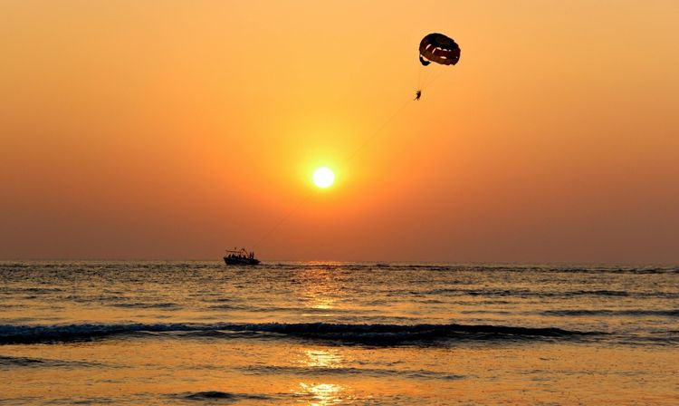 Silhouette of a parasailer and tow boat over a golden-orange ocean sunset, gentle waves reflecting the sun