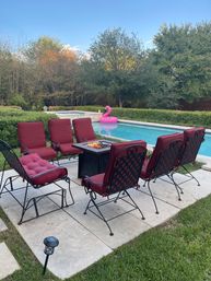 Backyard pool patio with six red-cushioned iron chairs arranged around a lit square fire pit on stone pavers, a blue swimming pool with a pink inflatable flamingo, and leafy trees under a clear sky.