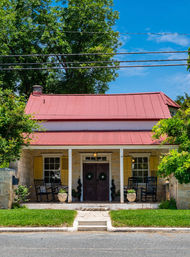Quaint historic stone cottage with a red metal roof, yellow shutters, shaded front porch with rocking chairs and potted plants, green lawn and bright blue sky.