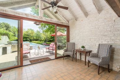 Sunny sunroom with vaulted whitewashed ceiling and ceiling fan, stone accent wall, two striped armchairs and wooden side table, sliding glass doors opening to a backyard pool patio with red umbrella, patterned lounge chairs and outdoor grill.