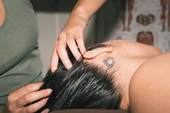 Close-up of a scalp massage: therapist hands massaging a person’s wet hair near a small heart-shaped neck tattoo and gold ear hoops, anatomy chart in the background.
