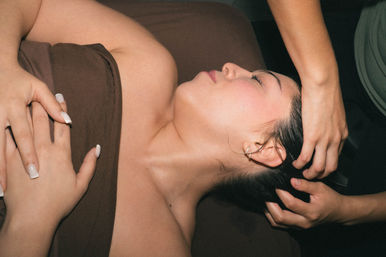 Serene woman wrapped in a brown towel lying on a spa table with eyes closed while a therapist massages her scalp, hands resting on her chest.