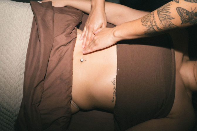 Close-up overhead shot of a person receiving a gentle abdominal massage on brown linens, hands on the midriff, visible navel piercing and floral forearm tattoo.